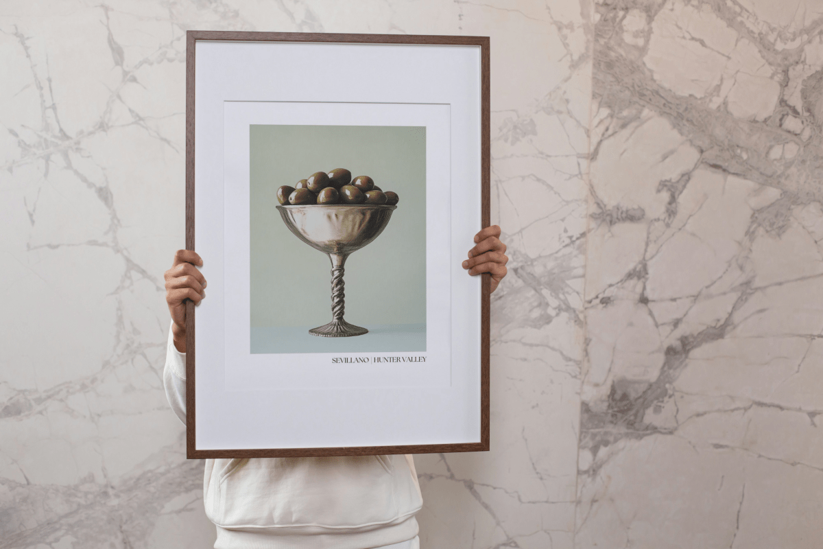 Person holding a framed Thirsty Merchy Sevillano olive poster against a marble wall. Featuring a metallic goblet filled with olives, this elegant artwork celebrates Hunter Valley’s gourmet culture and complements Stay n' Sip's premium wine-tasting experiences. Available for purchase online