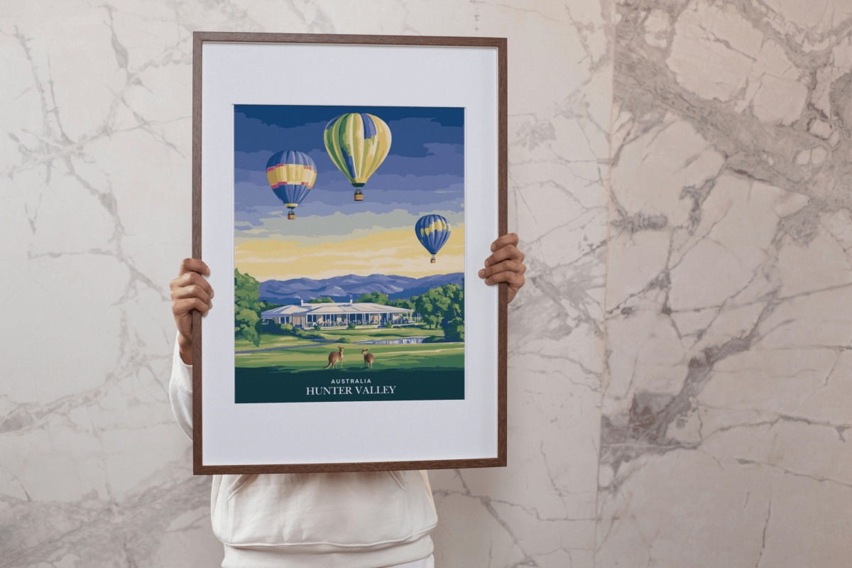 A framed art print featuring vibrant hot air balloons soaring over the Hunter Valley landscape, with kangaroos grazing and a grand estate in the backdrop. The artwork is elegantly held by a person, showcasing its size and rich colors, ideal for home or office decor