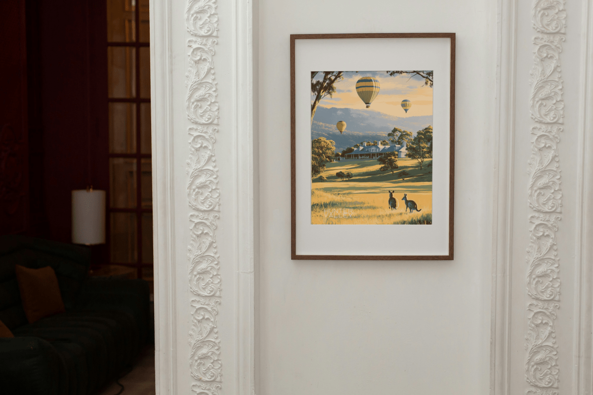 Framed Thirsty Merchy poster depicting a serene Hunter Valley landscape with hot air balloons, a countryside estate, and kangaroos, displayed on an elegant white wall with ornate detailing. Part of Stay n' Sip's exclusive art collection celebrating Australian wine country, available for purchase online