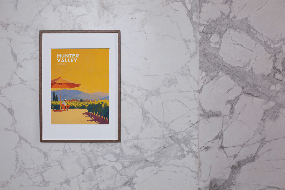 Framed Thirsty Merchy poster of a vibrant Hunter Valley vineyard scene, featuring a person relaxing under a red umbrella against a golden sky. Displayed on a marble wall, this artwork is part of Stay n' Sip's exclusive collection, celebrating Australian wine country and available for purchase online