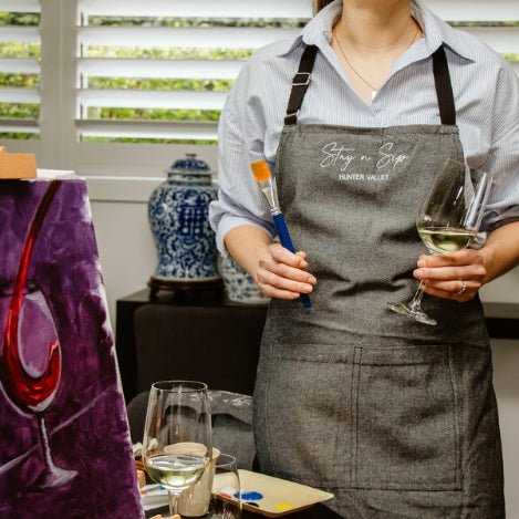 Person in a Stay n' Sip branded apron holding a wine glass and a paint brush.