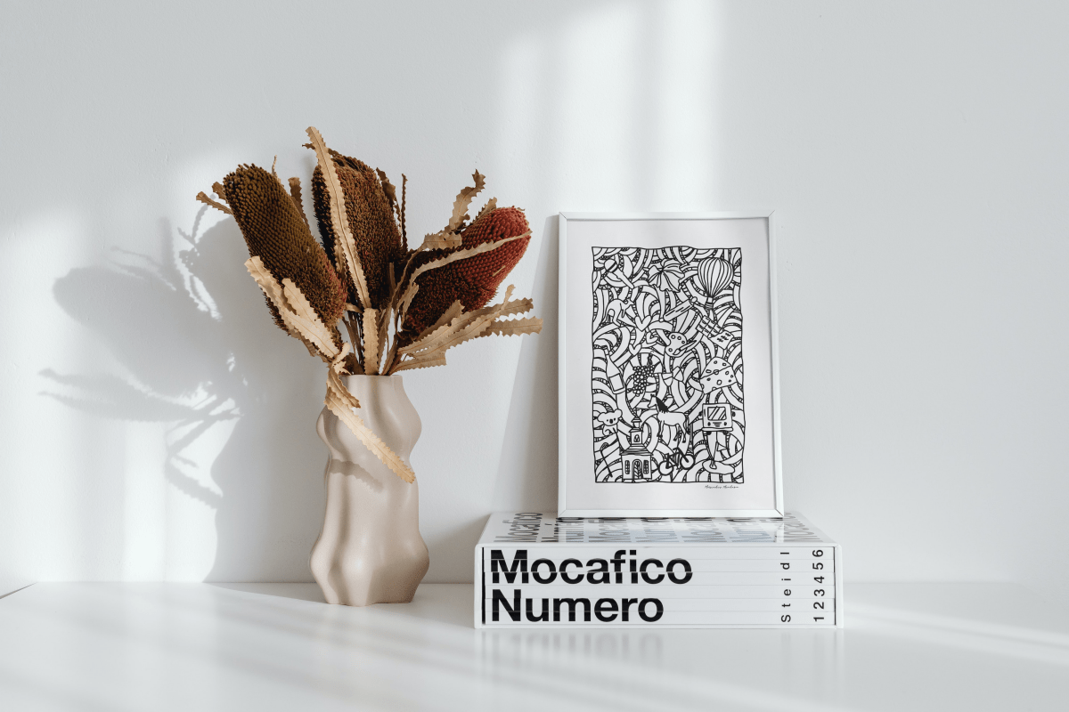 Framed Hunter Valley Doodle Poster displayed on a minimalist white surface, leaning against a wall. The black and white doodle design features whimsical elements representing the Hunter Valley, including hot air balloons, vineyards, and local wildlife. The setup is complemented by a modern, sculptural vase with dried native foliage, casting artistic shadows on the wall, and a stack of stylish coffee table books, creating a contemporary and artistic ambiance