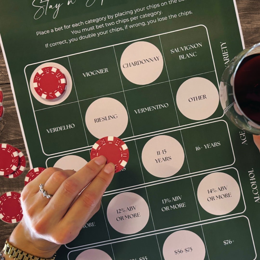 A close-up of Stay n' Sip's interactive wine tasting game board, featuring wine varieties, age, and alcohol content categories. A hand with a diamond engagement ring is placing a red poker chip on the board, surrounded by more chips. A glass of red wine rests on the board's edge, highlighting the playful and engaging nature of the new wine tasting experience