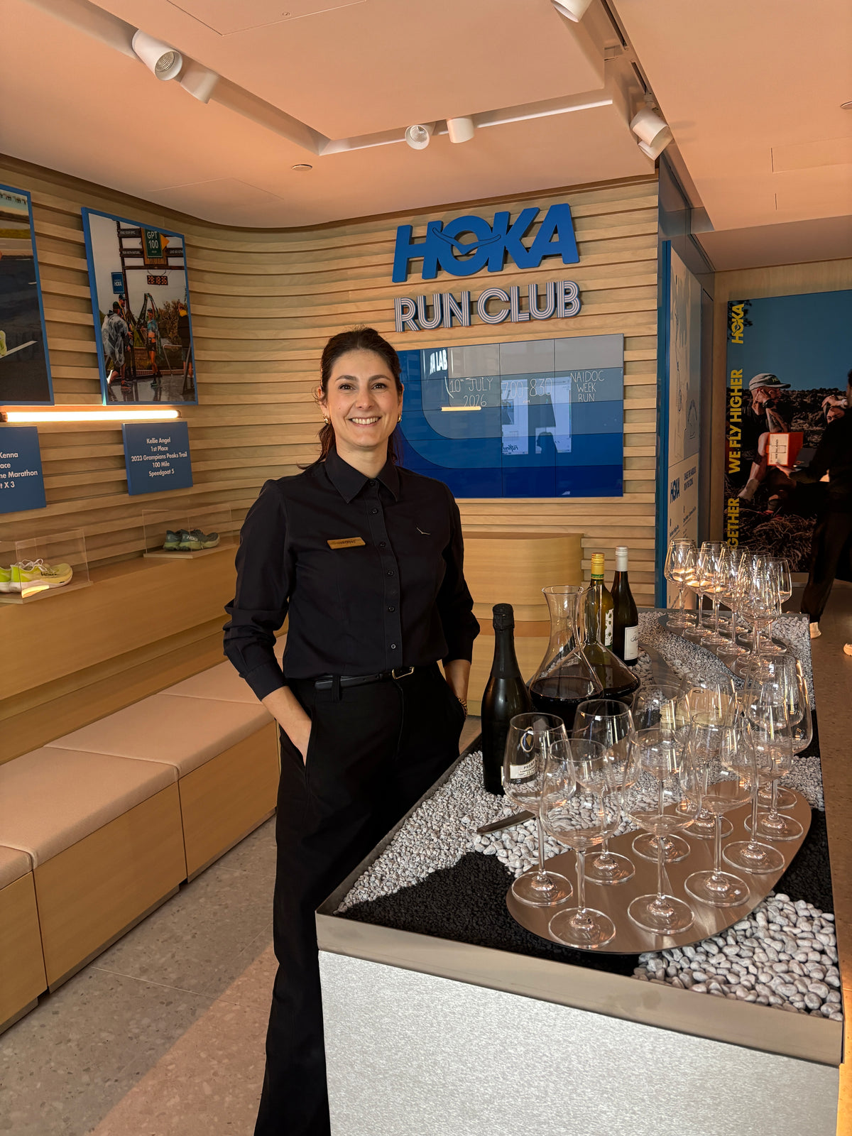 A woman in a black Hoka uniform stands behind a display counter at the Hoka Run Club, surrounded by wine bottles, glassware, and branded wall signage.