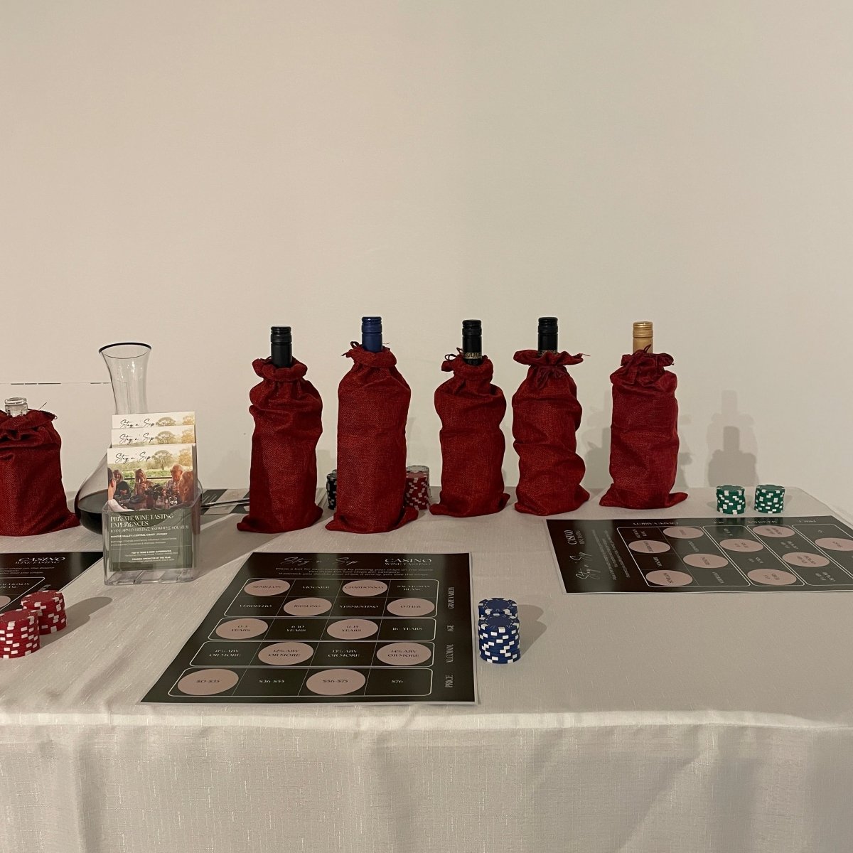 A sophisticated setup for Stay n' Sip's Casino Wine Tasting experience. Six wine bottles are elegantly concealed in deep red fabric bags, lined up on a white tablecloth. In front of the bottles, two Casino Wine Tasting game boards are displayed alongside stacks of red, blue, and green poker chips. A clear carafe and promotional brochures sit on the left, inviting guests to engage in this interactive and luxurious wine-tasting game that blends fun with wine education.