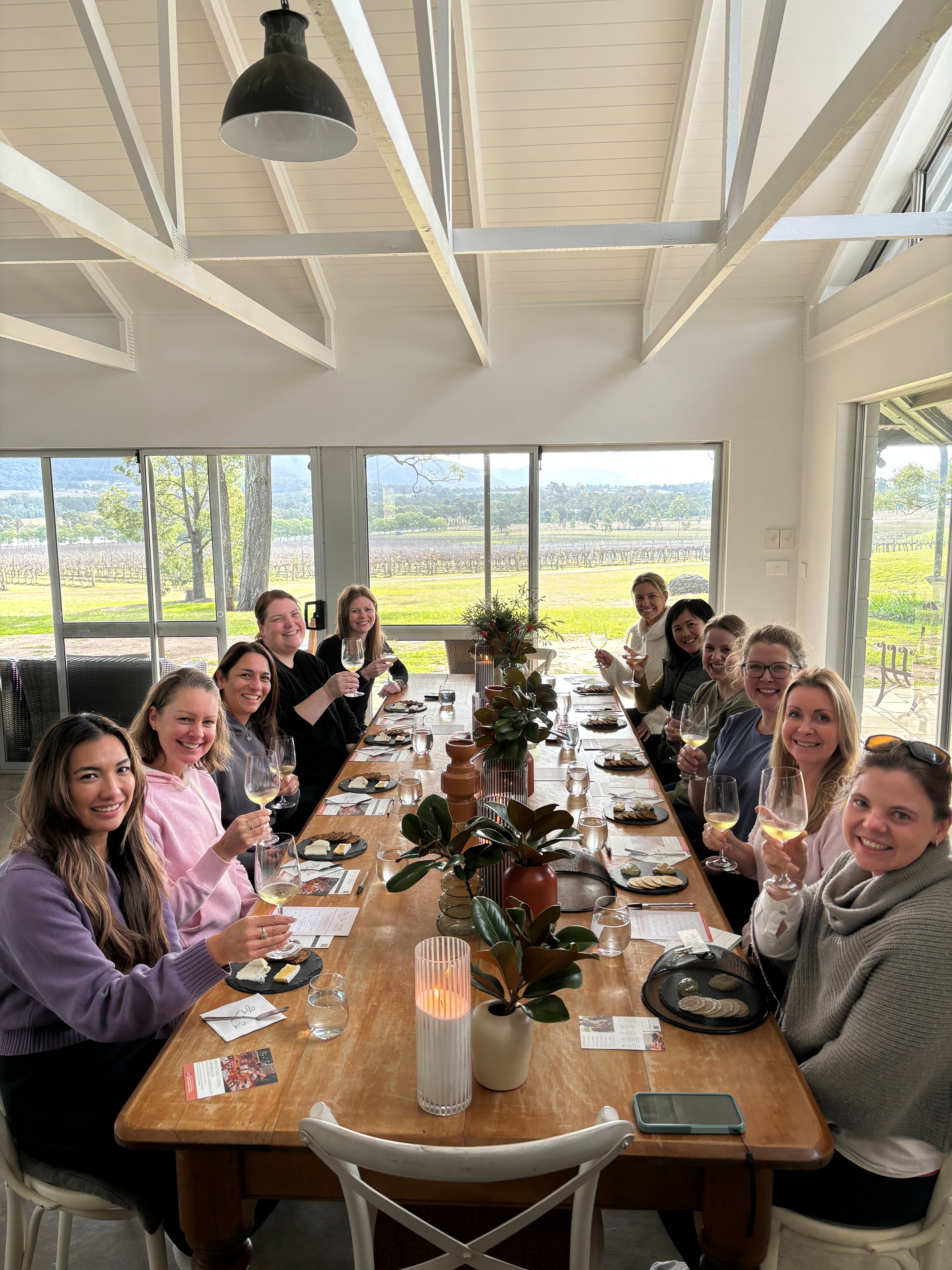 A group of 12 people enjoying a wine tasting experience, seated around a long wooden table in a bright room with large windows overlooking scenic vineyard views. The participants are smiling, holding glasses of wine, and engaging in conversation, with plates of cheese and snacks in front of them