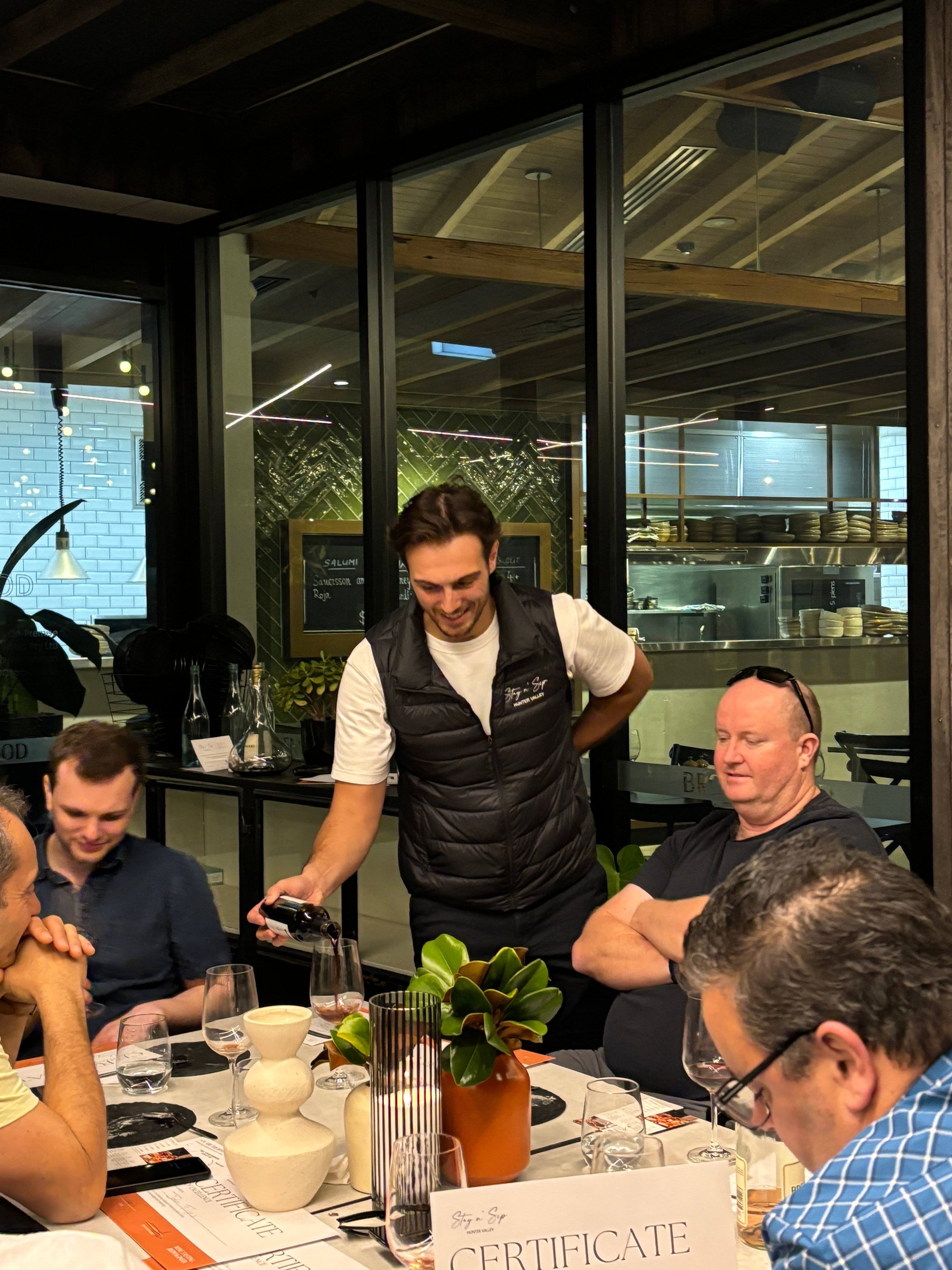 A wine educator from Stay n' Sip pours red wine for a group of participants during a Hunter Valley group wine tasting. The setting is indoors at a stylish venue, with guests engaged in conversation around a table adorned with tasting notes, wine glasses, and decorative vases