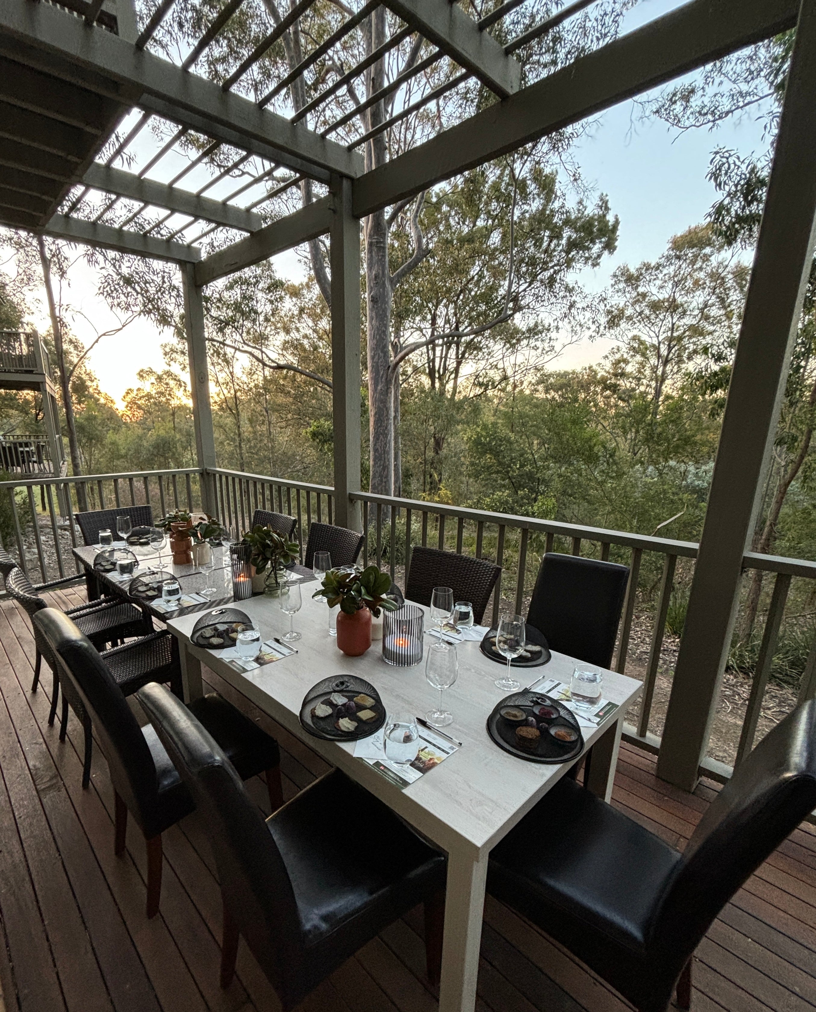 An elegant outdoor dining setup for a Hunter Valley group wine tasting on a deck surrounded by tall trees and nature. The long table is set with wine glasses, tasting plates covered with cloches, and decorative vases, ready for guests to enjoy the experience in the peaceful, natural setting at sunset.