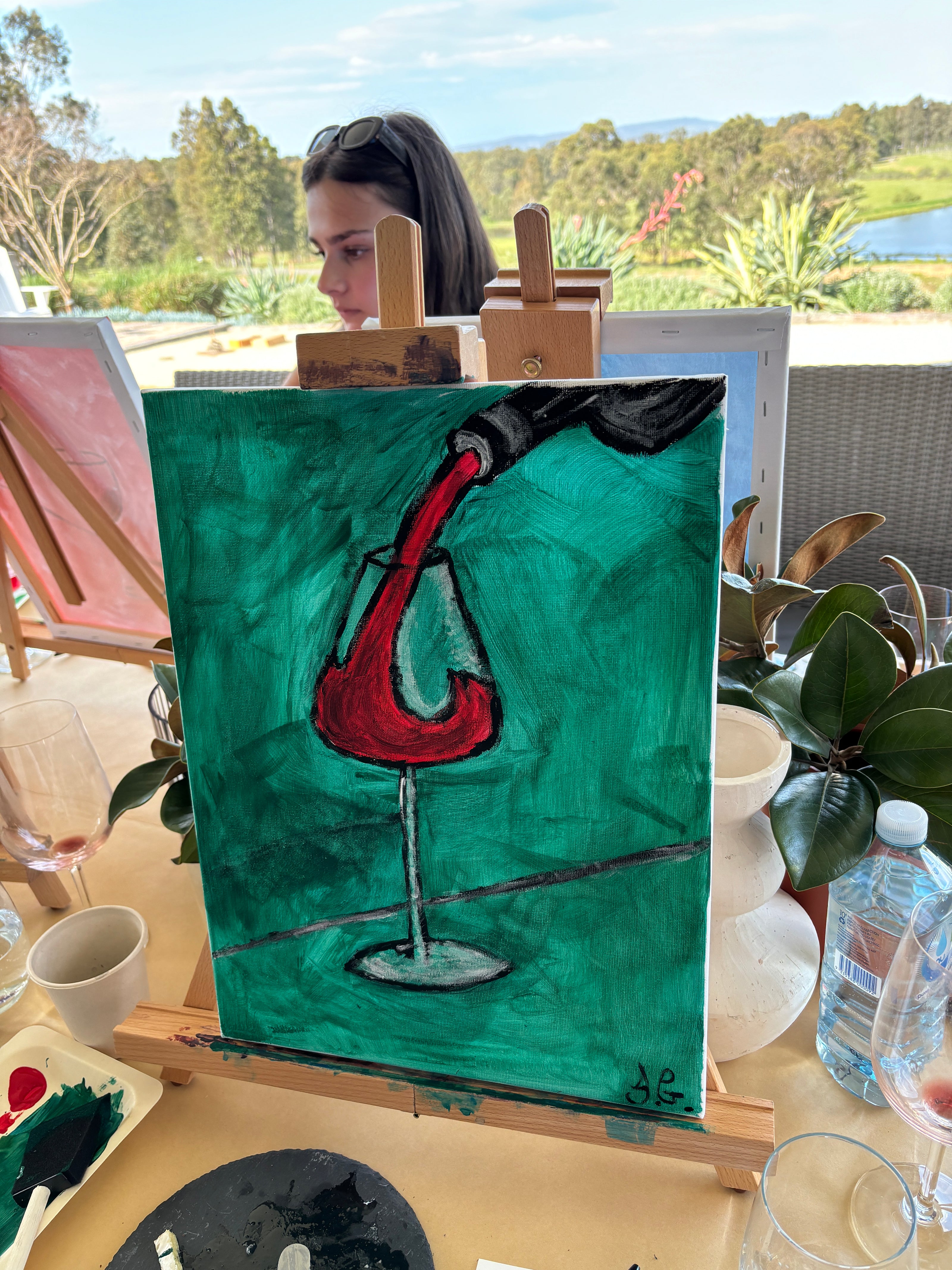 A painting of red wine being poured into a glass on an easel during a Hunter Valley group wine tasting and painting experience. The setting is outdoors with a scenic backdrop of greenery and a lake, while participants work on their individual canvases