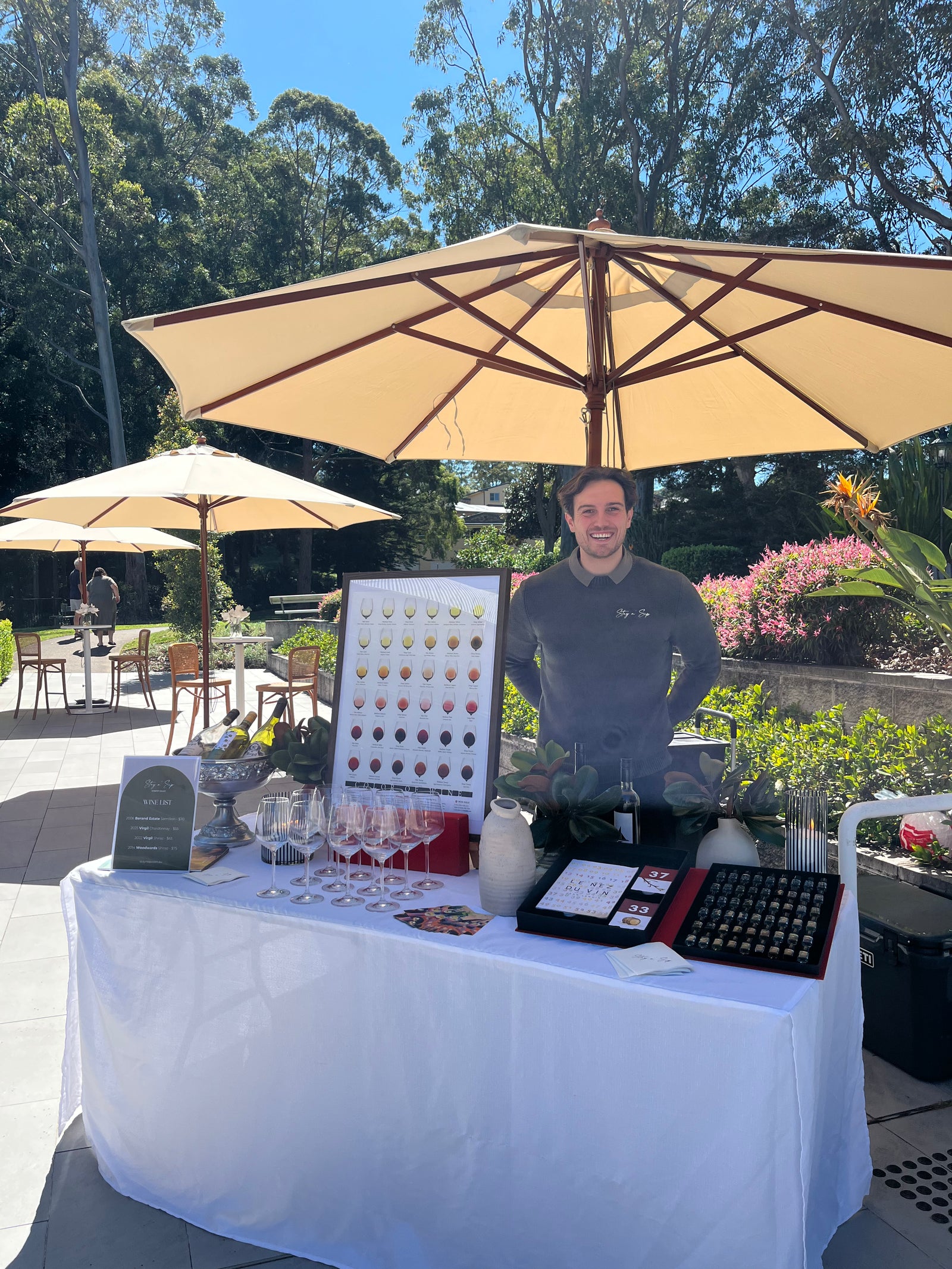 A man stands behind a white-covered table at an outdoor wine tasting event, under large beige umbrellas. The table holds a display board with many small circular aroma samples (Le Nez du Vin 54 Aromas), glasses, cards, and a posters with different wine colours nuances.