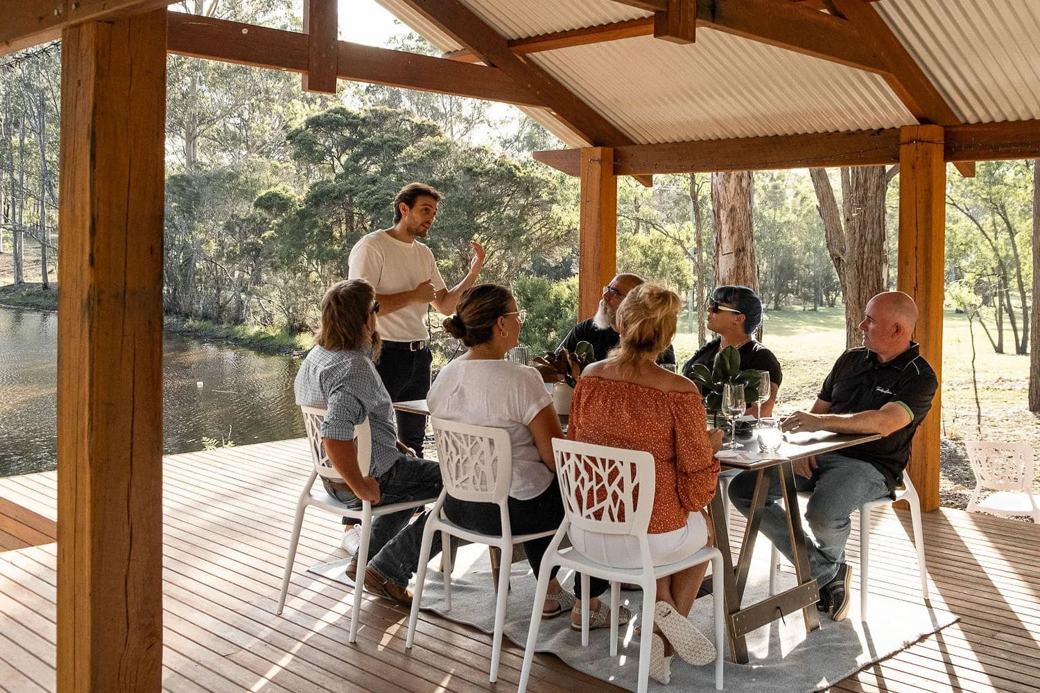 Group of six adults enjoying a wine tasting experience in Australian bushland overlooking a dam.