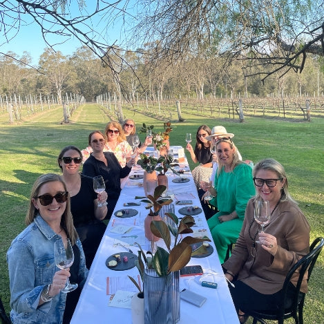 Long table with group of guests facing the camera with wine glasses and vineyard in the background.